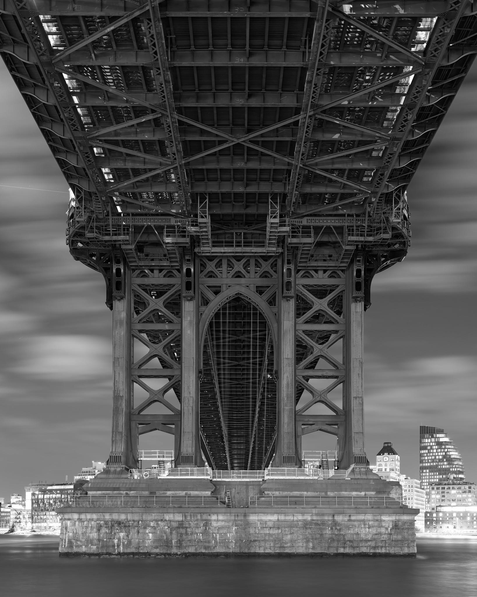 Underside of an early 20th century bridge, exposing girders and steelwork, over a river, at night.