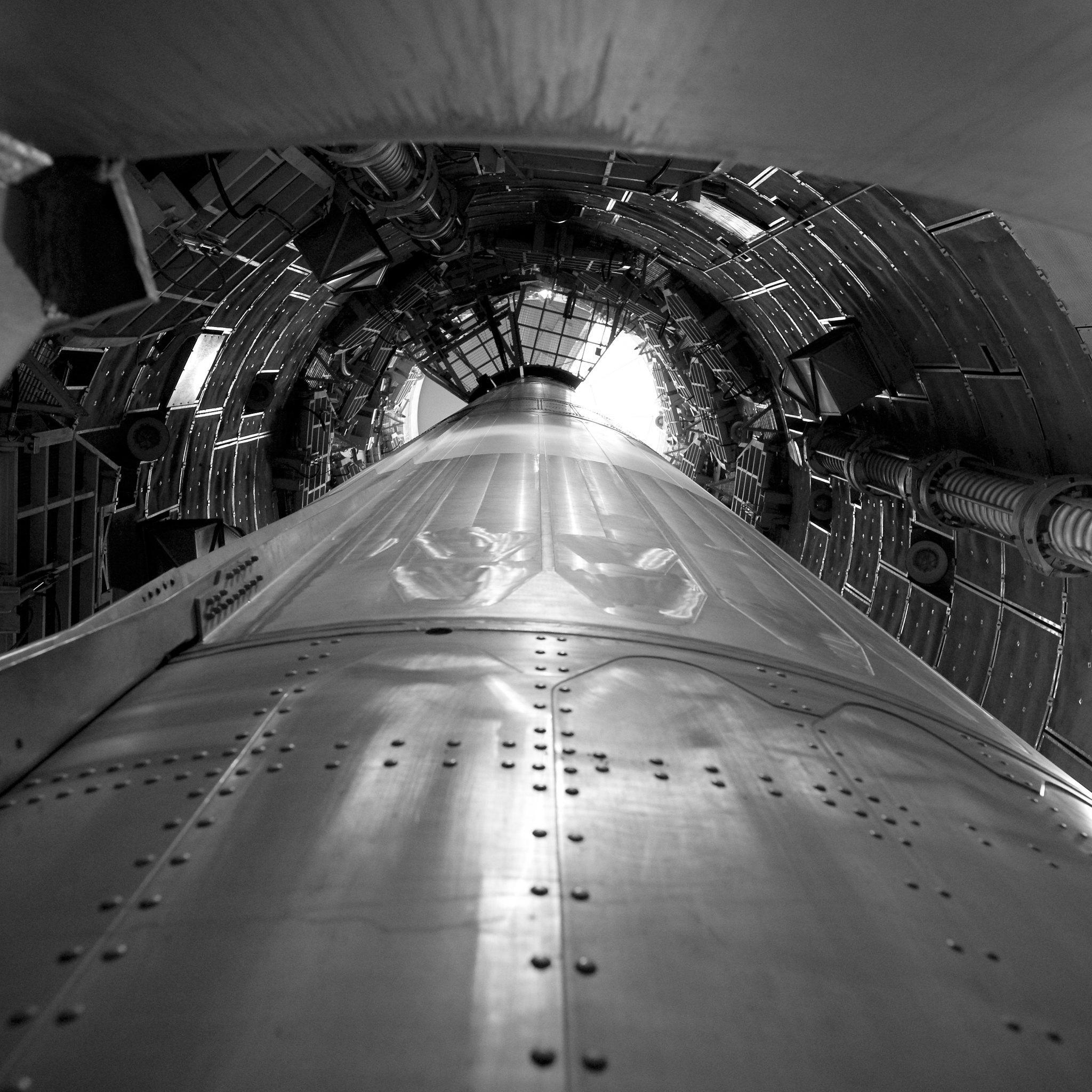 A shiny missile seen looking up from the bottom of a cramped underground silo, lined with platforms and various apparatus.