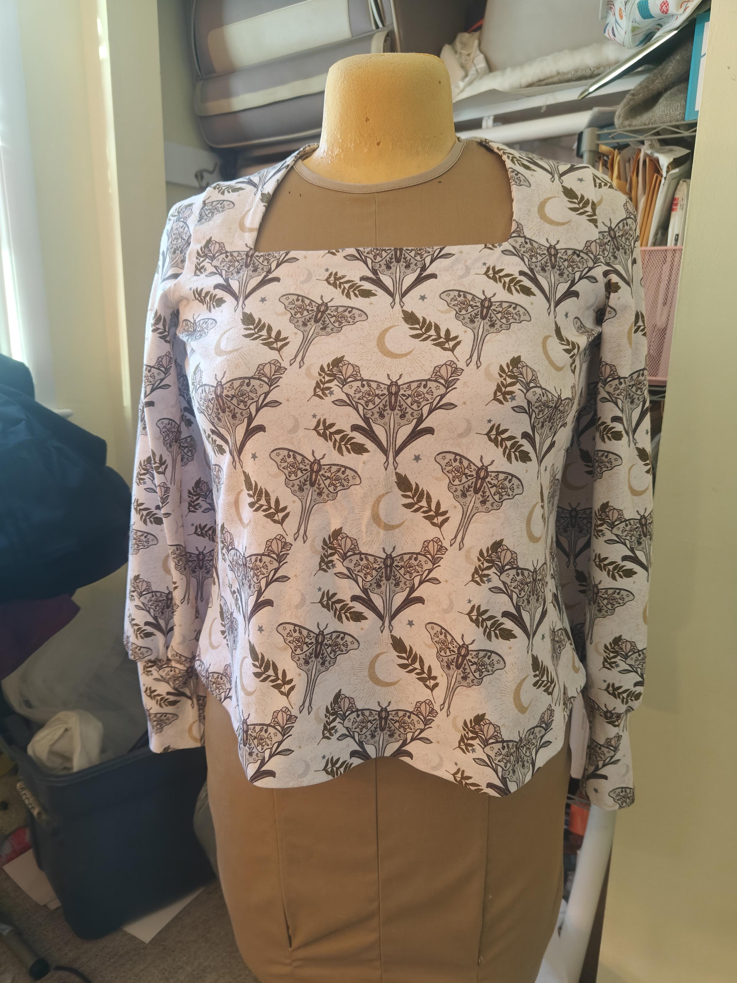 On a dressmaker's mannequin is a knit top with a Queen Anne neckline and bishop sleeve. The print is light sage green and lavender moths and gold crescent moons on a white background 